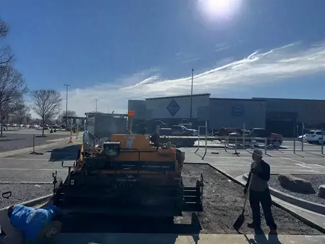 Paving crew and loaded truck working across a large retail lot