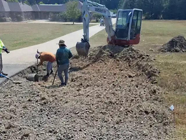 Mini excavator and crew moving soil beside a paved path